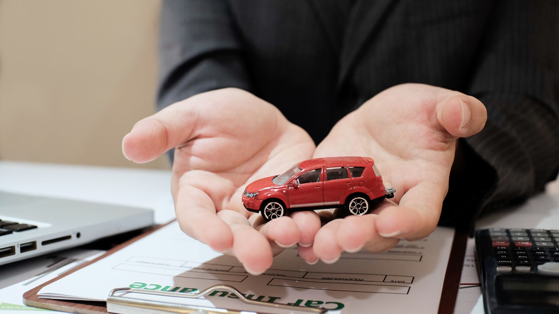 Hands holding a small red toy car over insurance paperwork and calculator